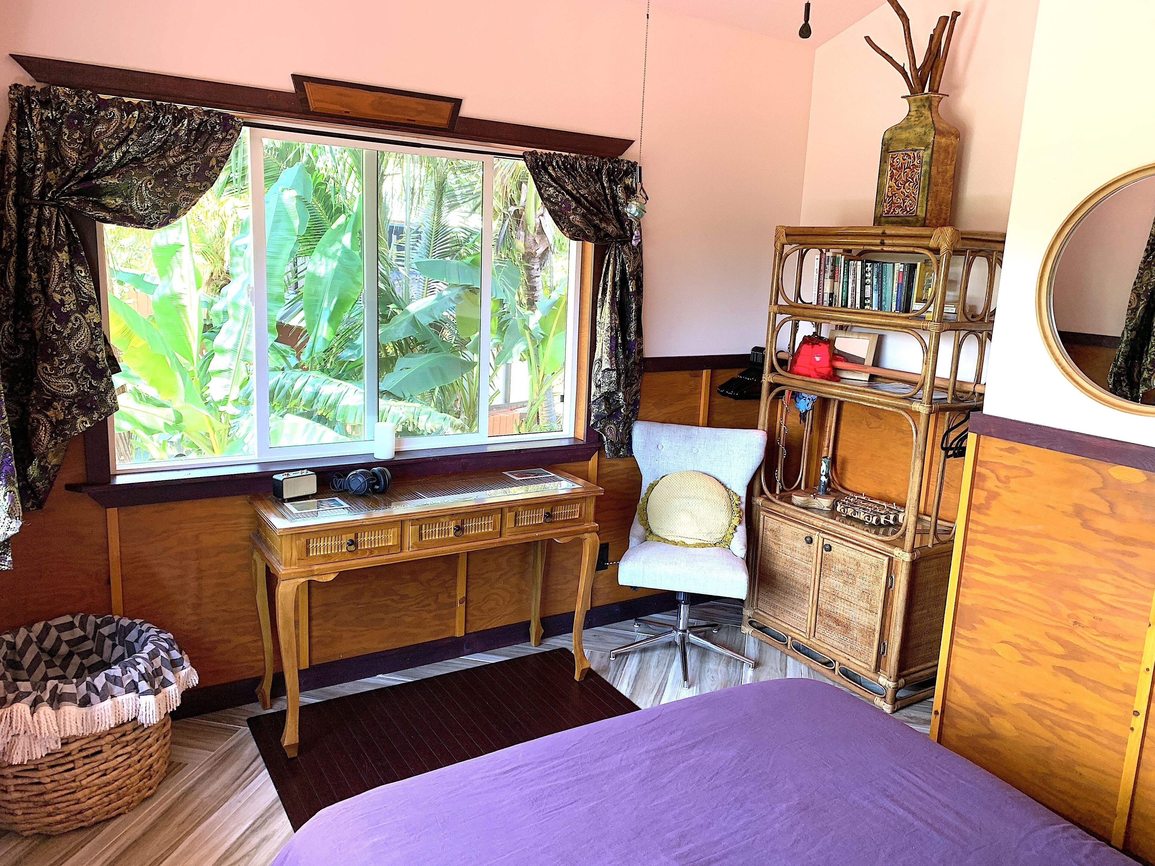 Una habitación con un ventanal grande, un escritorio de madera, una silla, una estantería y una cama con colcha púrpura.