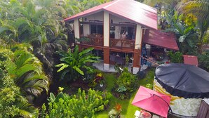 Uma casa de dois andares com telhado vermelho, cercada por vegetação tropical exuberante.