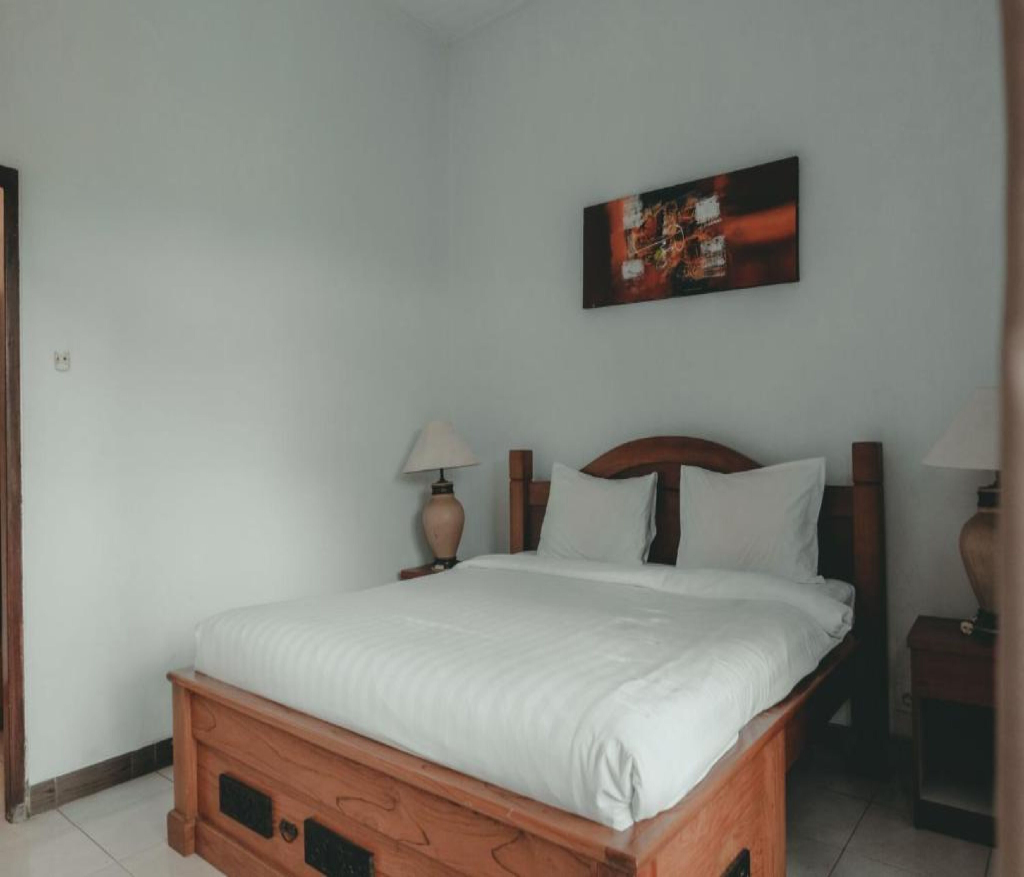 Un lit bien fait, avec du linge de lit blanc, dans une pièce aux murs blancs. On y trouve une tête de lit en bois et deux tables de chevet avec des lampes. Un tableau est accroché au mur, au-dessus du lit.