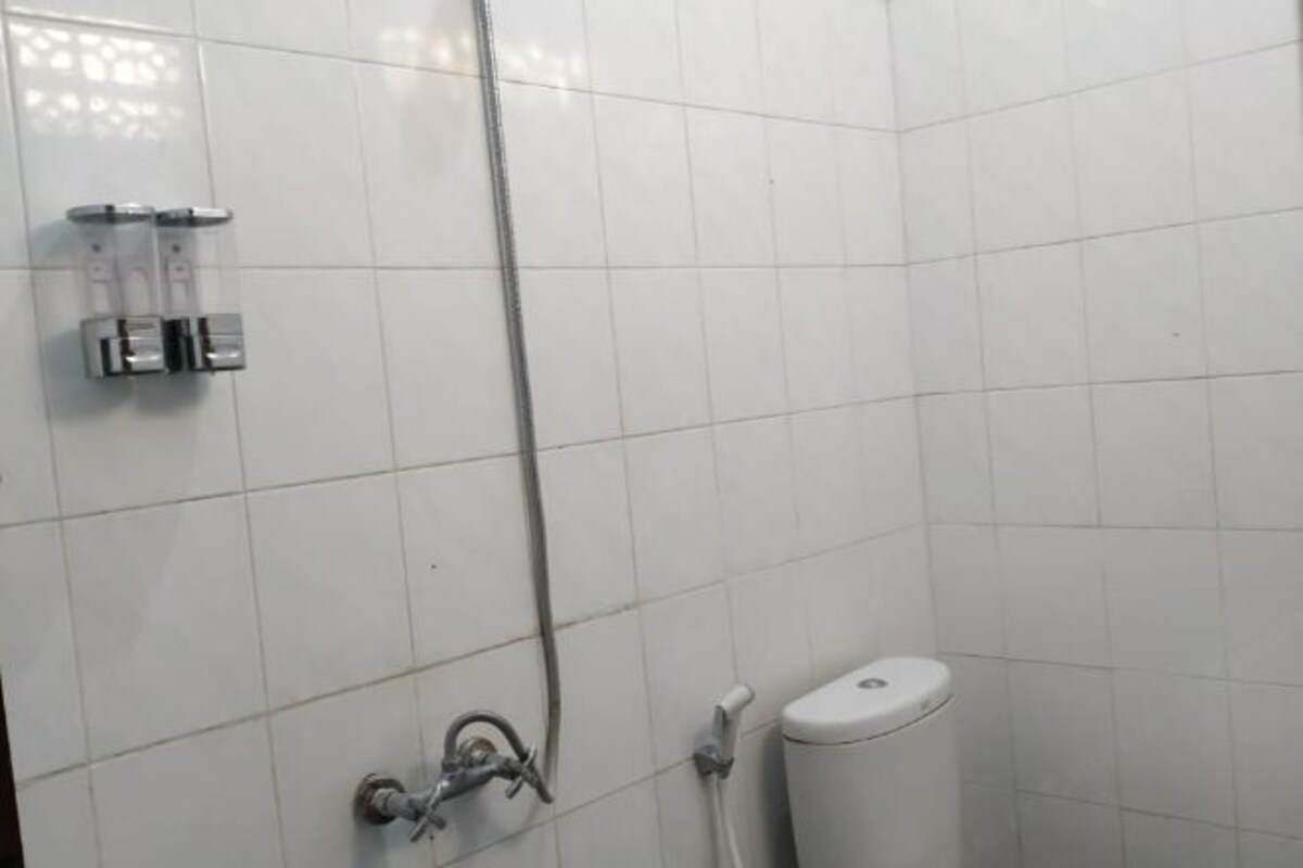 Ett badrum med vita kakelväggar, en dusch med ett justerbart duschmunstycke, en toalett och en väggmonterad tvåldispenser.
