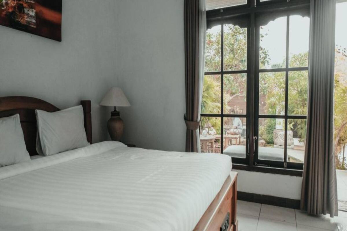 Una cama bien hecha con cabecera de madera, una lámpara de noche y vistas al exterior a través de ventanas grandes.