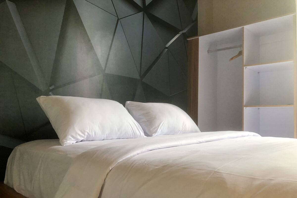 Un dormitorio moderno con un cabecero de estampado geométrico, una cama bien tendida con ropa blanca y un armario minimalista con acabado de madera.