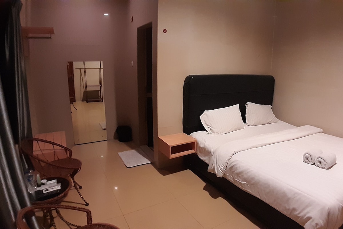 Habitación de hotel ordenada con una cama individual, una mesita de noche, una silla y una pequeña mesa con una botella y un mando a distancia.