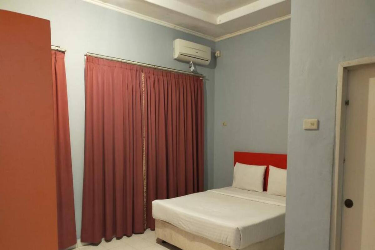 A small, single-bed room with a red headboard, a window with red curtains, and an air conditioning unit mounted on the wall.