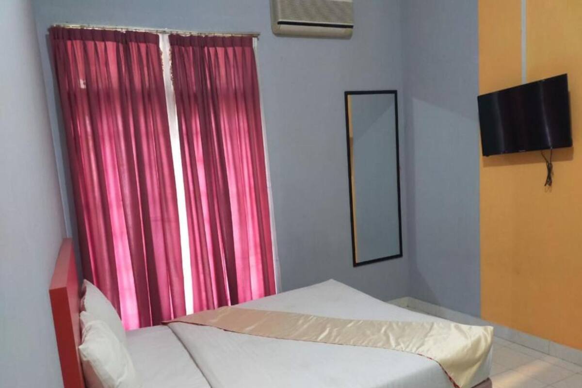A single bed with a white mattress and a beige blanket, pink curtains, a wall-mounted TV, and a small air conditioning unit.