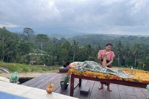 Una mesa de masajes al aire libre, con una persona acostada sobre ella, otra persona de pie a un lado, y un paisaje exuberante al fondo.
