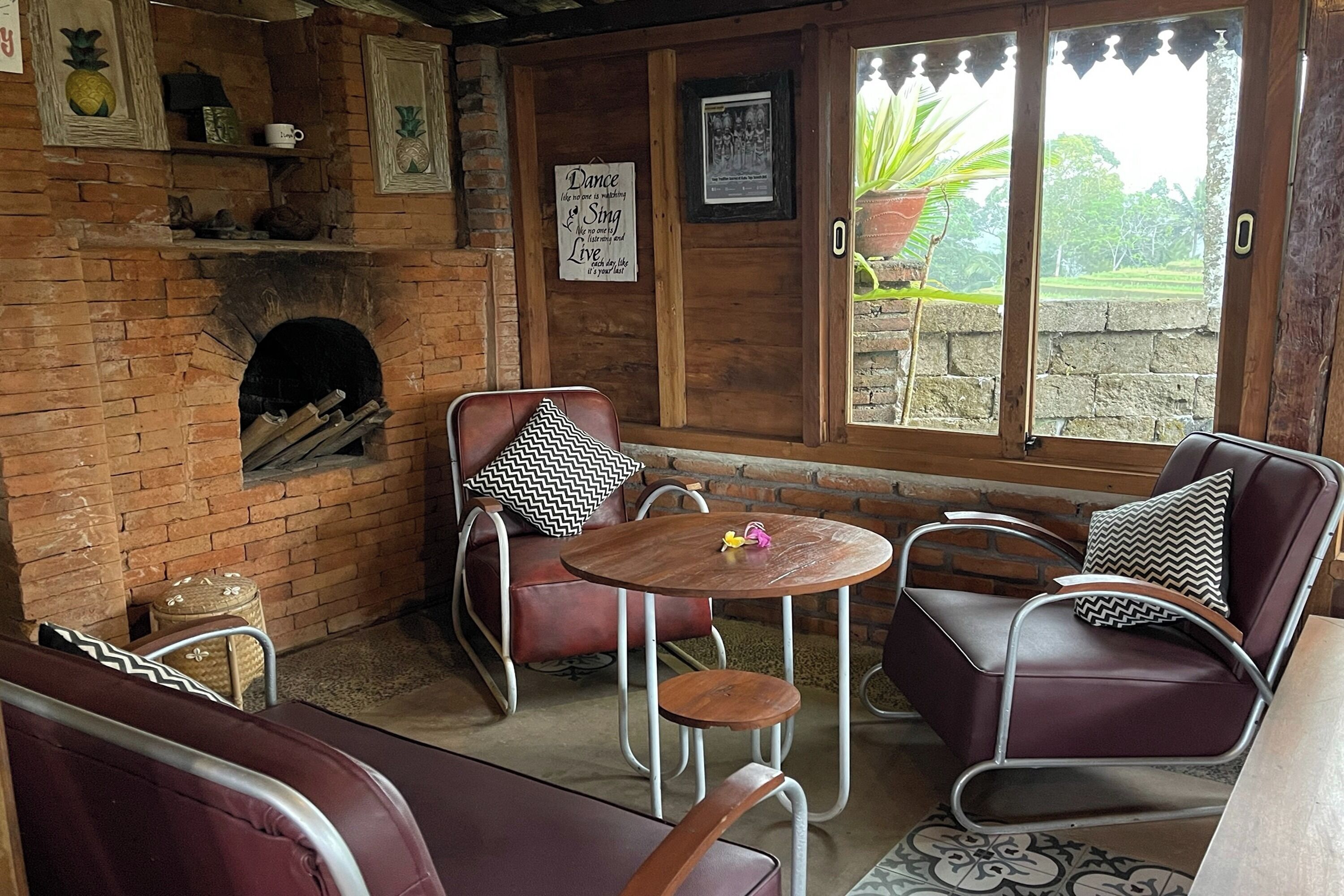 A cozy room with a brick fireplace, wooden walls, and a large window with a view of greenery. There are leather sofas and a small round table.