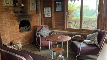 A cozy room with a brick fireplace, wooden walls, and a large window with a view of greenery. There are leather sofas and a small round table.