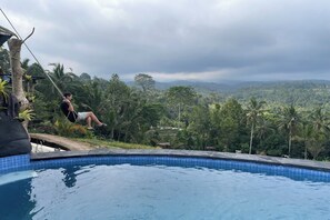 Una piscina con vistas a un paisaje exuberante y una persona balanceándose en una cuerda.