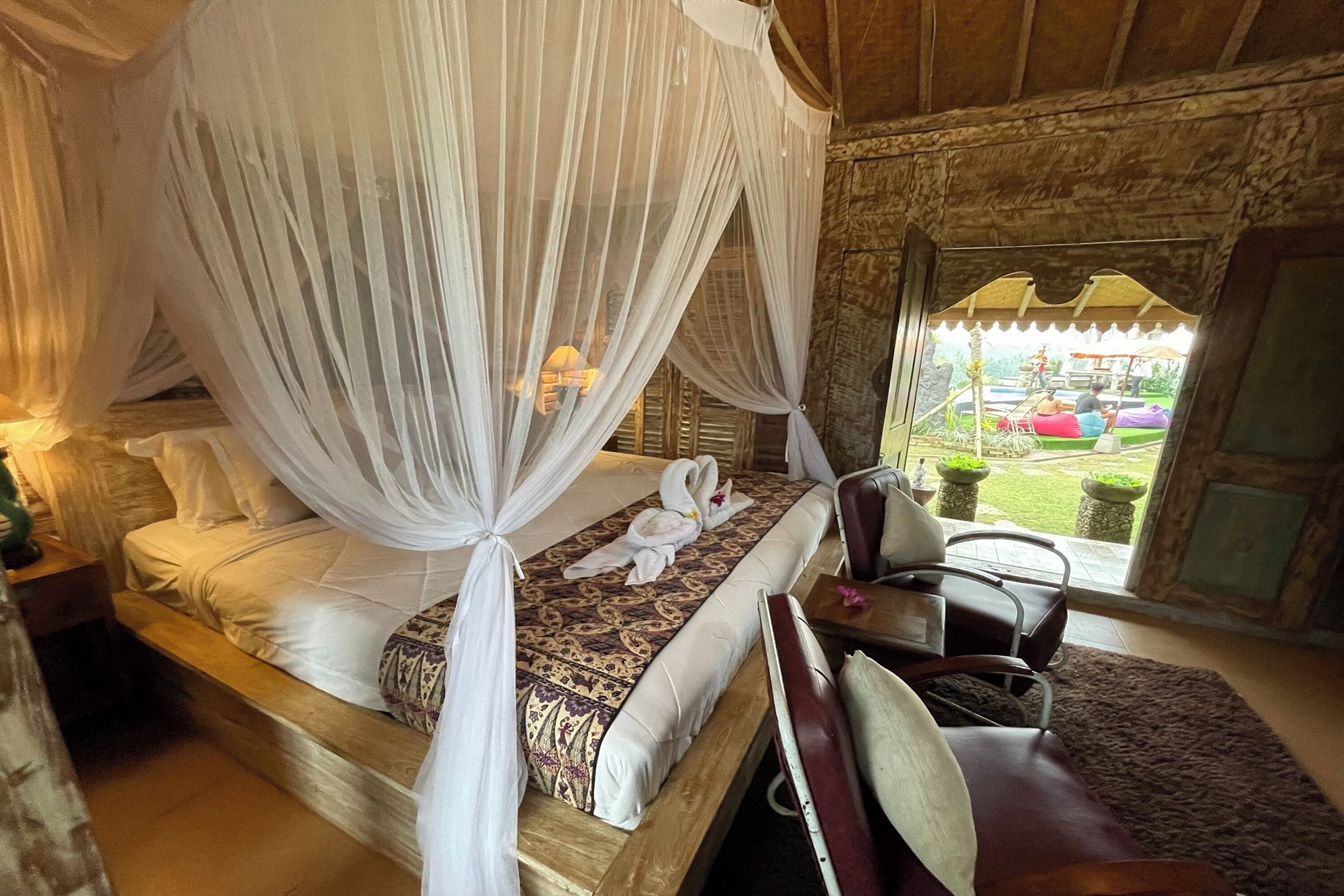A room with a bed under a canopy, a wooden headboard, a small table with a lamp, and a view of the outside.
