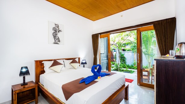Ein Hotelzimmer mit Holzboden, einem Bett mit brauner Bettdecke, einer blau gestalteten Handtuchskulptur, einem Nachttisch mit Lampe und Blick auf Grünpflanzen durch die Glastür.