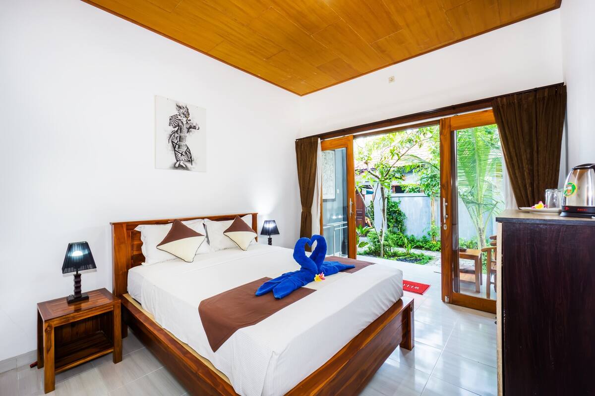 Une chambre d’hôtel avec un plafond en bois, un lit recouvert d’une couverture marron, une décoration en forme d’œuvre d’art réalisée avec une serviette bleue, une table de chevet avec une lampe, et une vue sur la verdure à travers la porte vitrée.