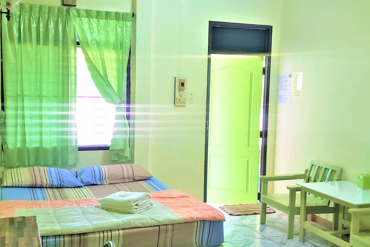 A room with a bed, a table, and a chair. There is a window with curtains, a door, and a wall-mounted television.