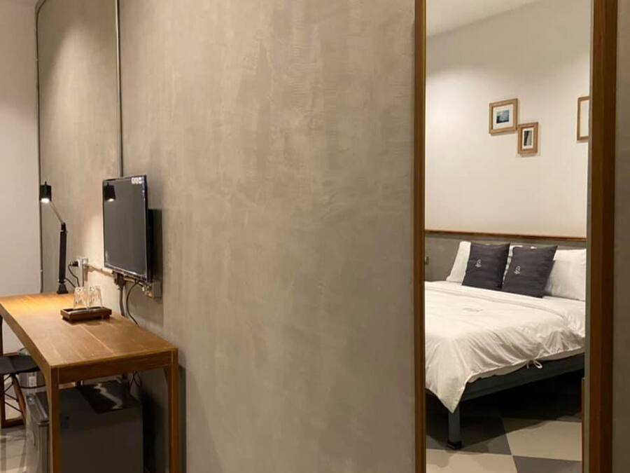 Quarto com cama, escrivaninha com computador e televisão na parede.