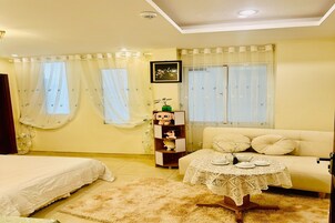 Una habitación con cama, sofá, una mesita con mantel de encaje y una estantería con objetos decorativos.