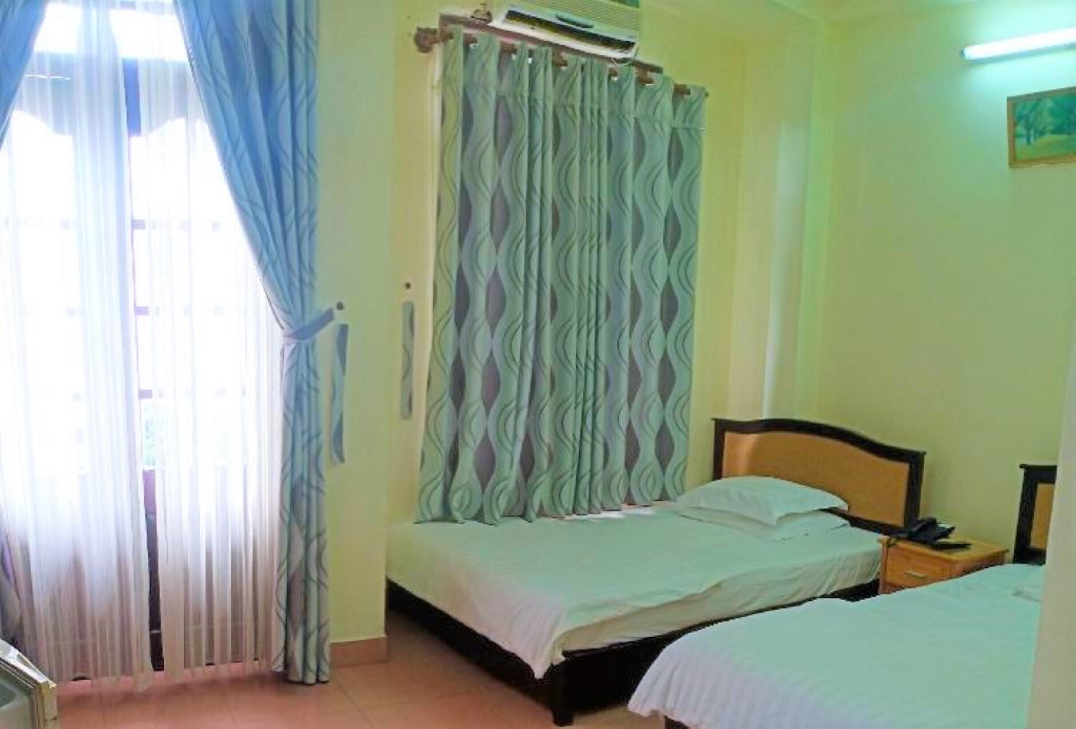 A room with two beds, a window with sheer curtains, an air conditioning unit, and a bedside table with a lamp.