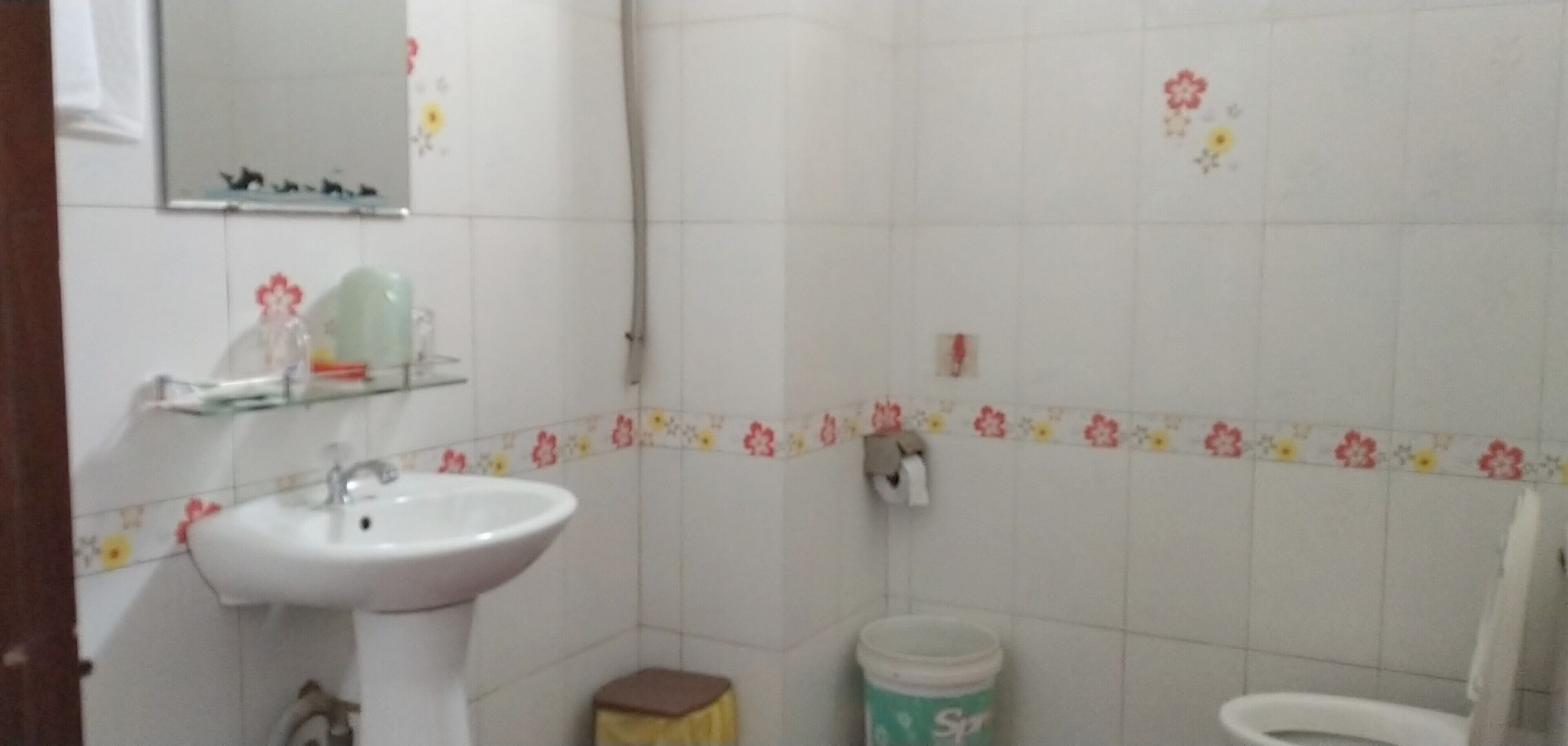 Une salle de bain avec un lavabo blanc, des toilettes et un petit tabouret. Il y a un miroir au-dessus du lavabo et un porte-rouleau de papier toilette au mur.