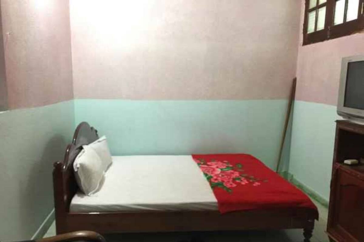 Une petite chambre avec un lit simple, un cadre de lit en bois, un téléviseur posé sur un meuble en bois et une couvre-lit à motifs floraux rouges.