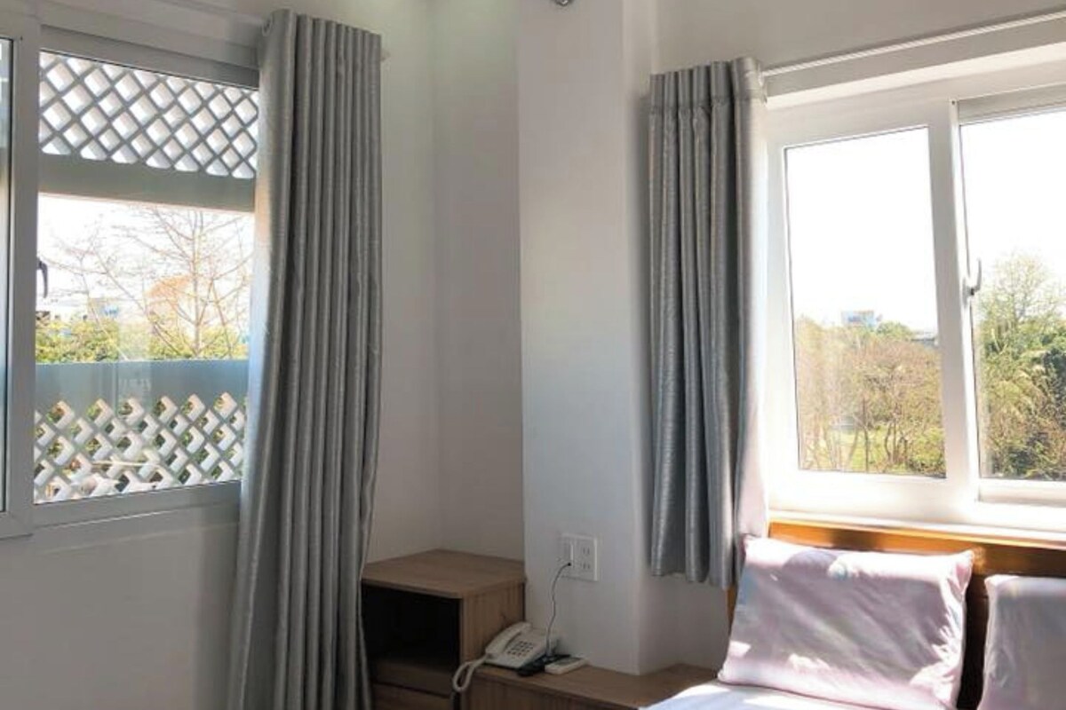 Une chambre d’hôtel avec un lit, une table de chevet avec un téléphone, une fenêtre avec des rideaux et une vue sur les arbres à l’extérieur.