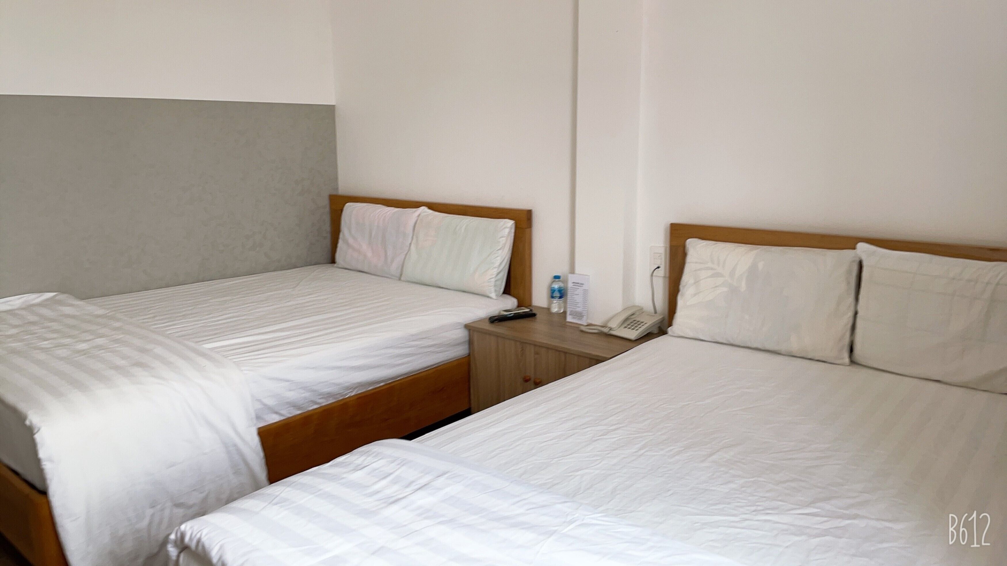 Twee enkele bedden met houten hoofdborden, een nachtkastje met een telefoon en een waterfles, en een effen muur.