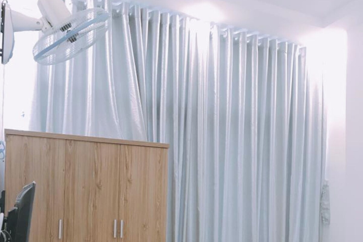 Une chambre avec une armoire en bois, un lit avec un matelas blanc, une petite table de chevet et un rideau.