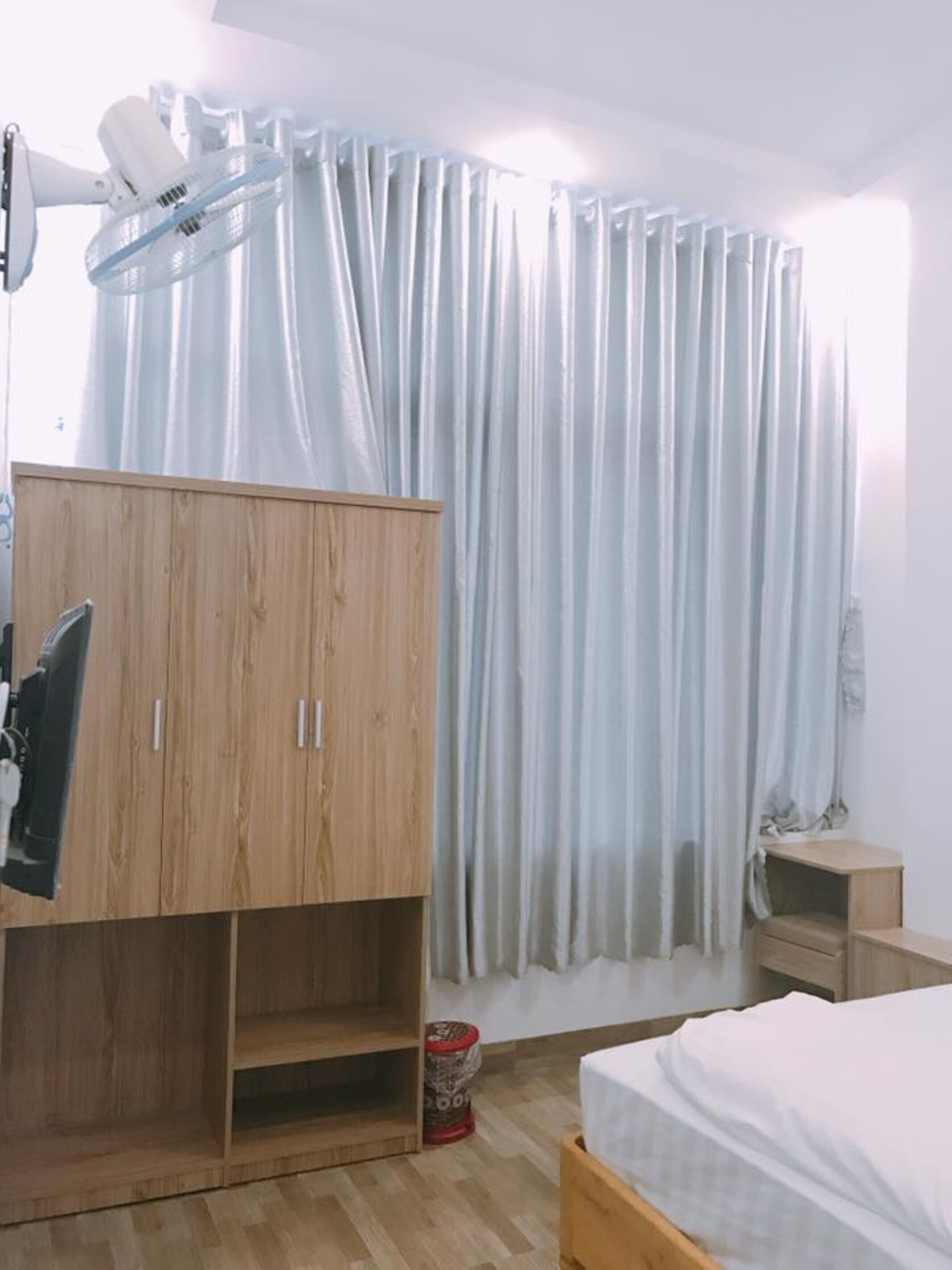 Une chambre avec une armoire en bois, un lit avec une literie blanche, une petite table de chevet et une fenêtre ornée de rideaux légers.