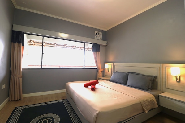 Una habitación de hotel con cama, una ventana con cortinas, un reloj de pared y una lámpara de noche.