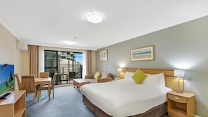 Ein Hotelzimmer mit einem Bett, Nachttischen, einem Schreibtisch mit Stuhl, einem Fernseher und einem Balkon mit Blick auf Gebäude.