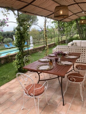 An outdoor patio with a table set for dining, surrounded by chairs with cushions, a pool in the background, and a covered area with a pergola.