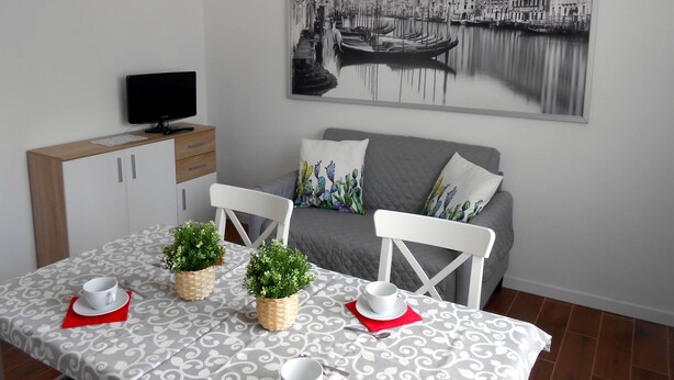 Een eetgedeelte met een tafel voor twee personen, een televisie op een houten kast en een stadsgezichtschilderij aan de muur.