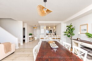 A modern dining area with a long wooden table, white chairs, and pendant lighting. - Dolphin Beach C110 Beachfront Beauty by hmh (Cape Town)