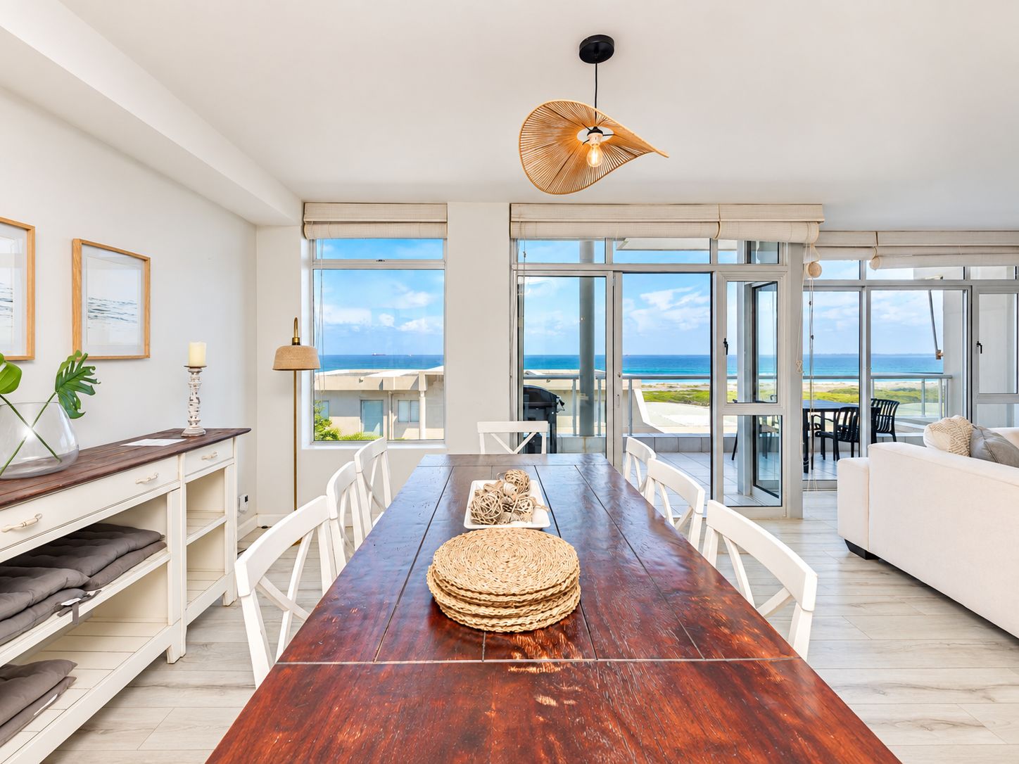 A spacious dining area with a wooden table, white chairs, and a view of the ocean through large glass doors.