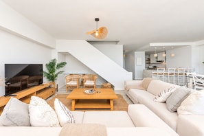 A modern living room with a flat-screen TV, a sofa set, a dining area with chairs, and a staircase leading to an upper level.