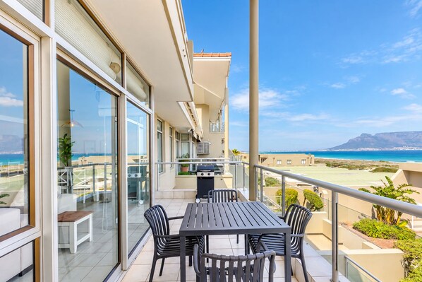 A balcony with a table and chairs, overlooking a beach and mountains.