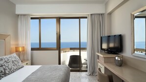 A hotel room with a bed, a TV, a desk, a chair, and a view of the sea through large windows. - Atlantica Oasis (Limassol)