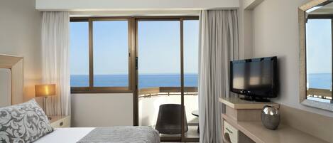 Habitación de hotel con cama, televisión, escritorio, silla y vistas al mar a través de amplias ventanas.
