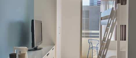 A modern, well-lit room with a flat-screen TV, a coffee maker, and a laptop on a white desk. A ladder leads to a balcony with a blue chair.