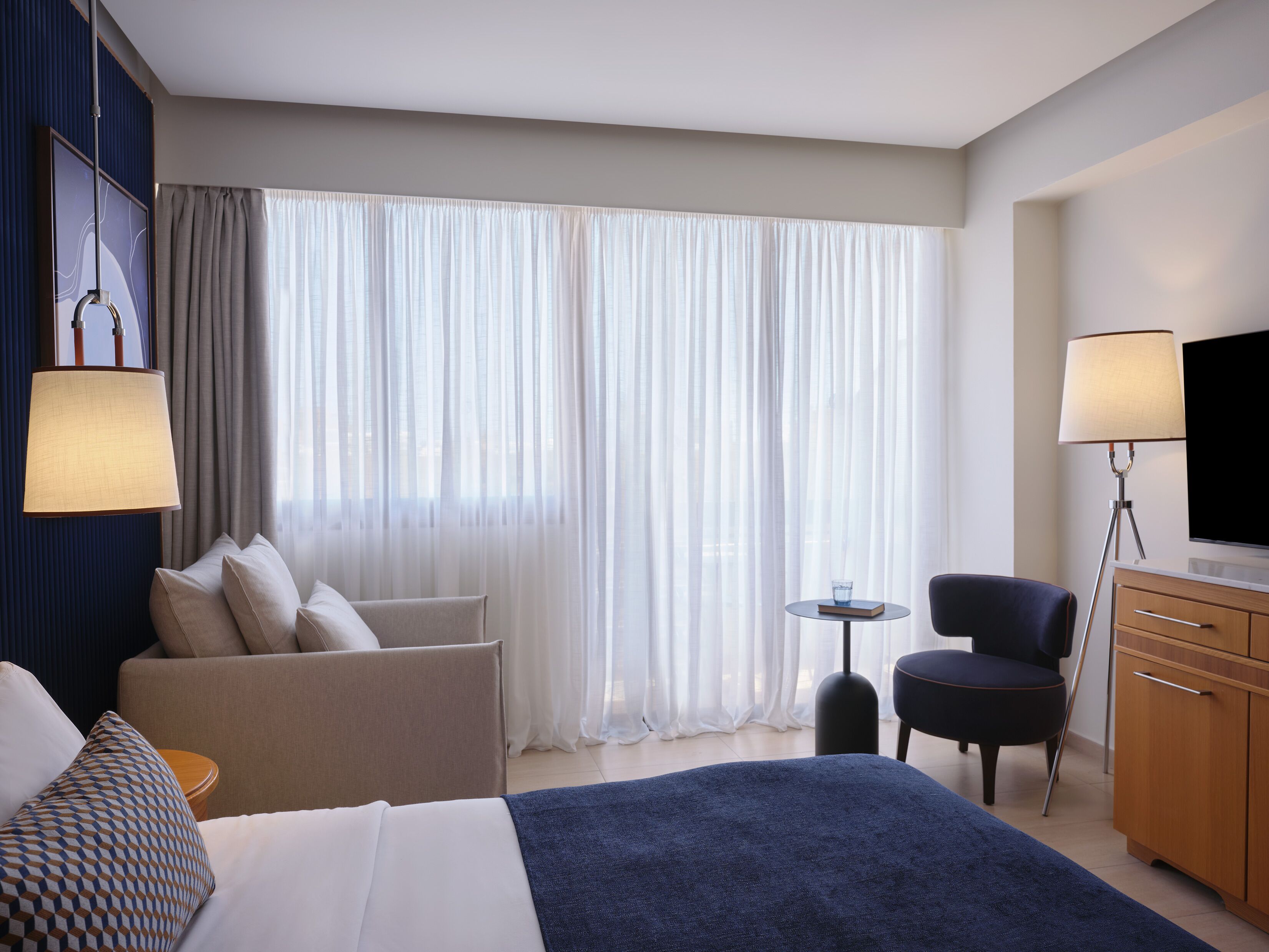 A hotel room with a bed, a sofa, a desk, a chair, a TV, and a lamp.