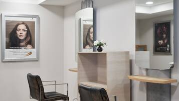 Een modern ingerichte kapsalon met twee kappersstoelen, een spiegel, een plank met een vaas, en ingelijste posters aan de muren.