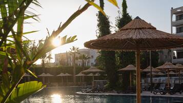 Zona junto a la piscina con sillones de descanso y sombrillas de paja, rodeada de palmeras y un cielo despejado.