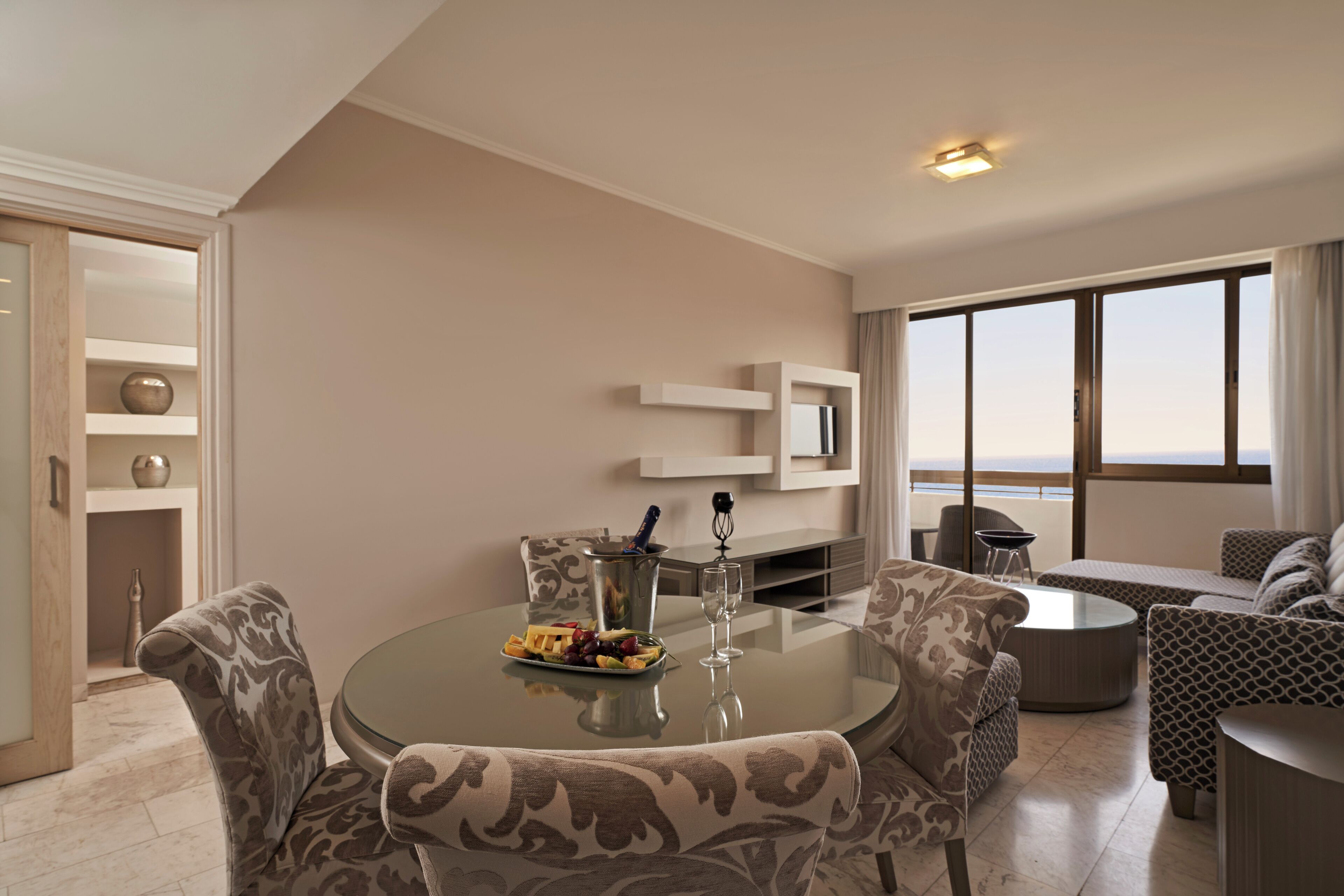 Una sala de estar moderna con una mesa de comedor de vidrio, sofás estampados y vistas al mar a través de amplios ventanales.