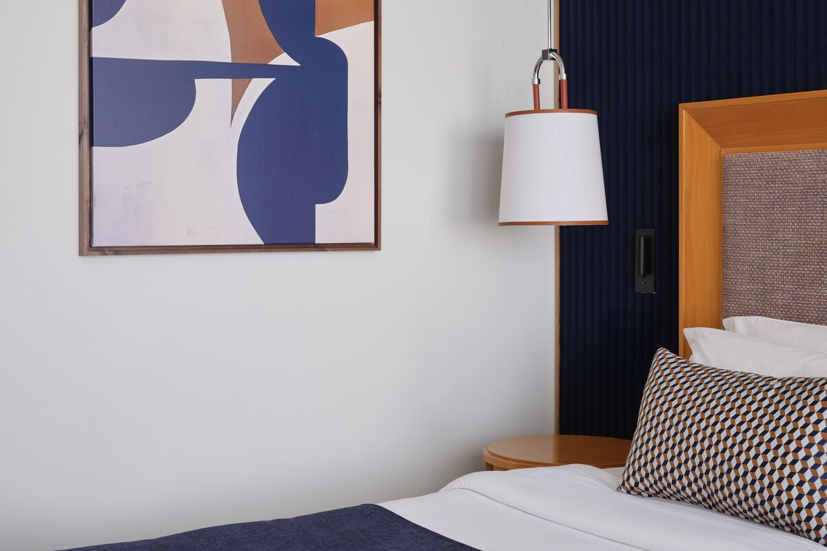 Una habitación de hotel con cama, mesita de noche, lámpara y un cuadro abstracto colgado en la pared.