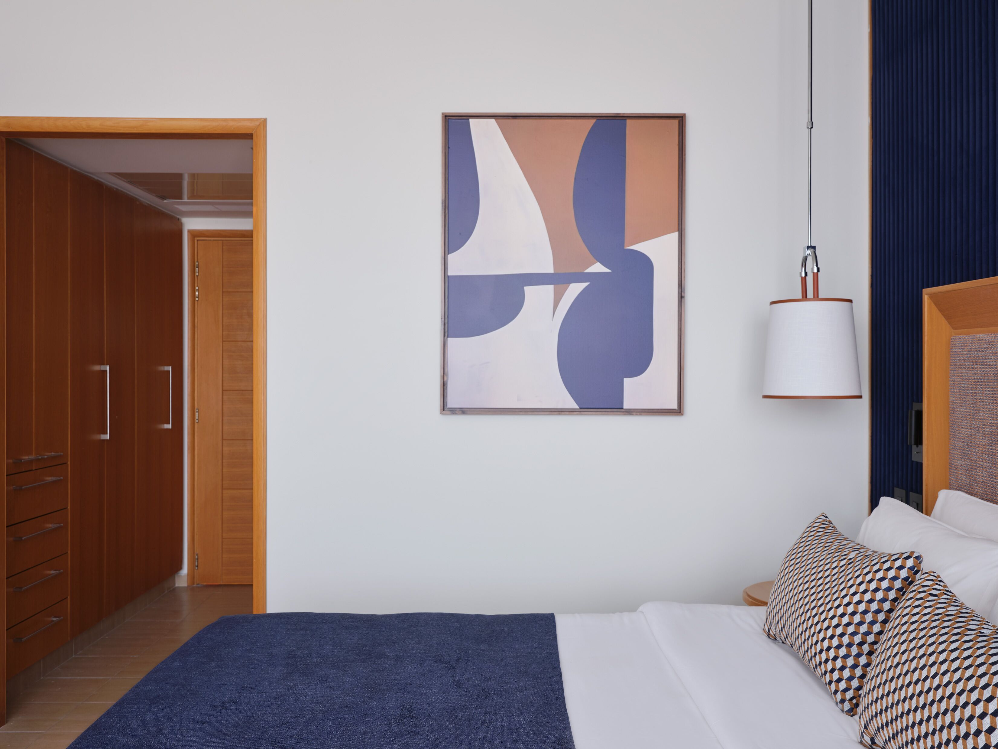 Une chambre à coucher avec un lit, une armoire en bois, un tableau abstrait encadré et une lampe suspendue.