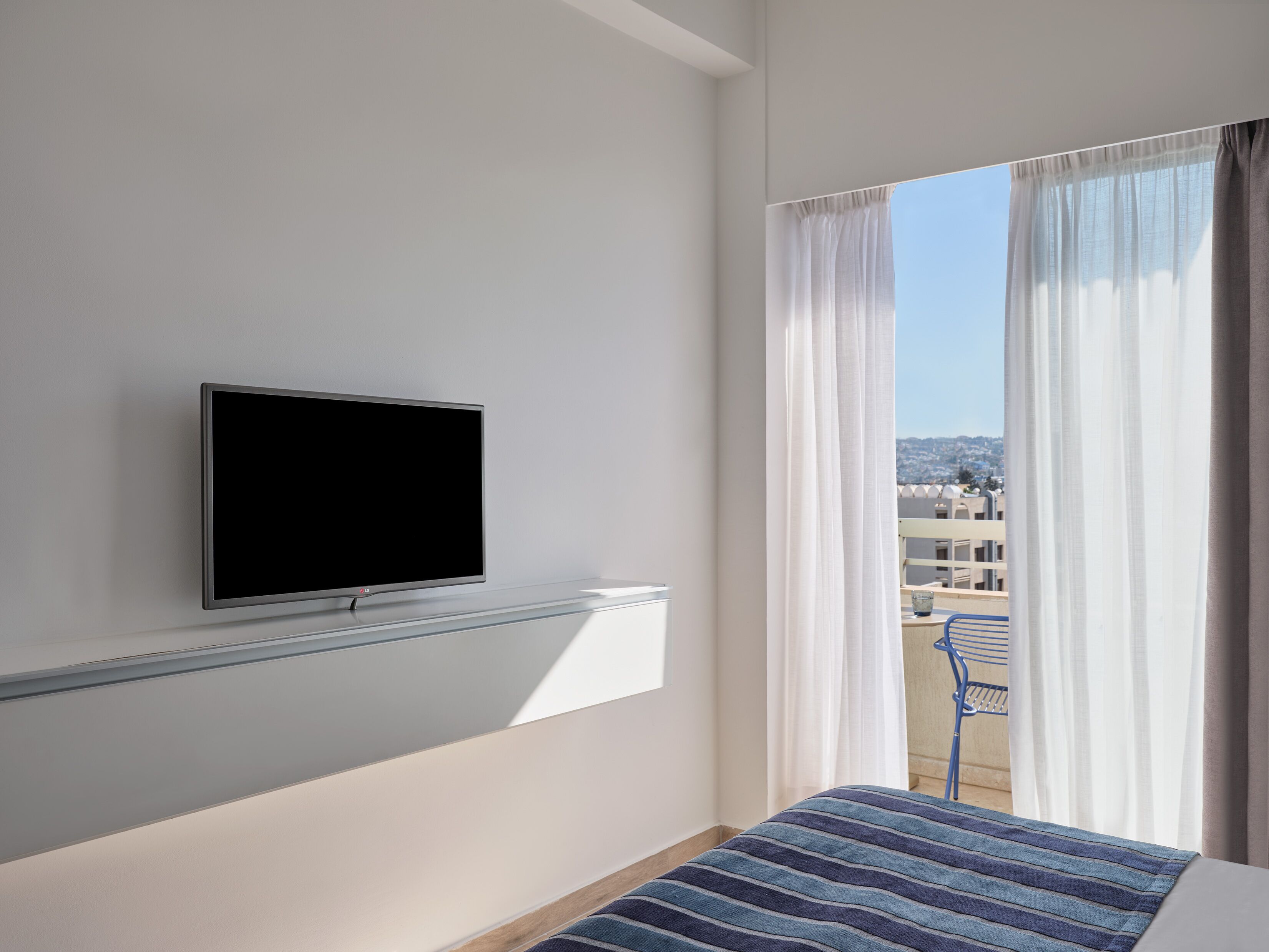 A hotel room with a mounted TV, a bed with a striped bedspread, a balcony with a view of buildings, and sheer curtains.