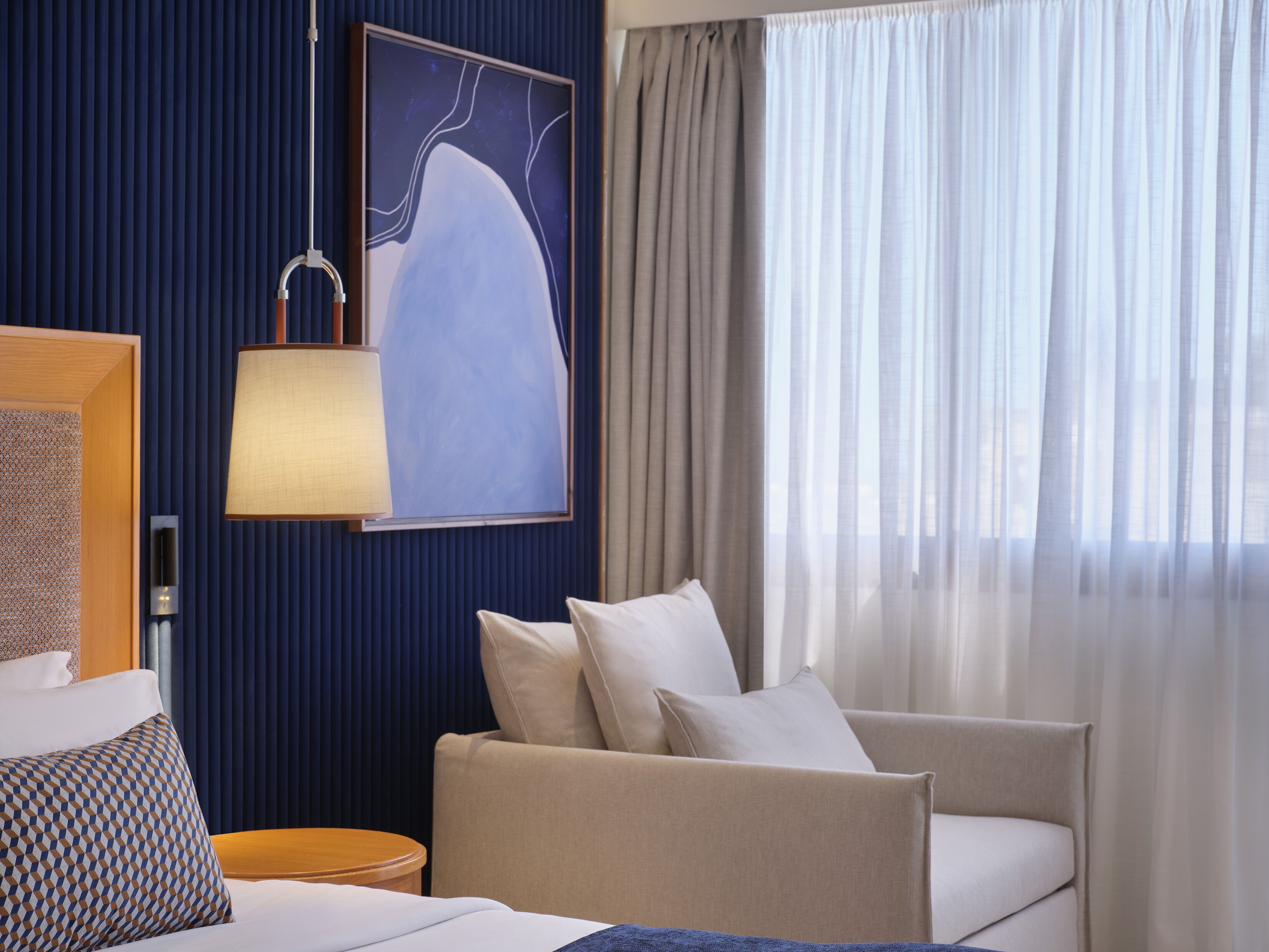 A hotel room with a bed, a sofa, a lamp, a painting, and a window with curtains.
