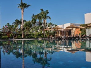 A pool with lounge chairs and umbrellas, surrounded by palm trees and a building with a stone facade. - Atlantica Oasis (Limassol)