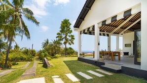 A modern outdoor patio with a dining area, palm trees, and a view of the ocean. - Villa Bloom by Villa Finder (Sukasada)