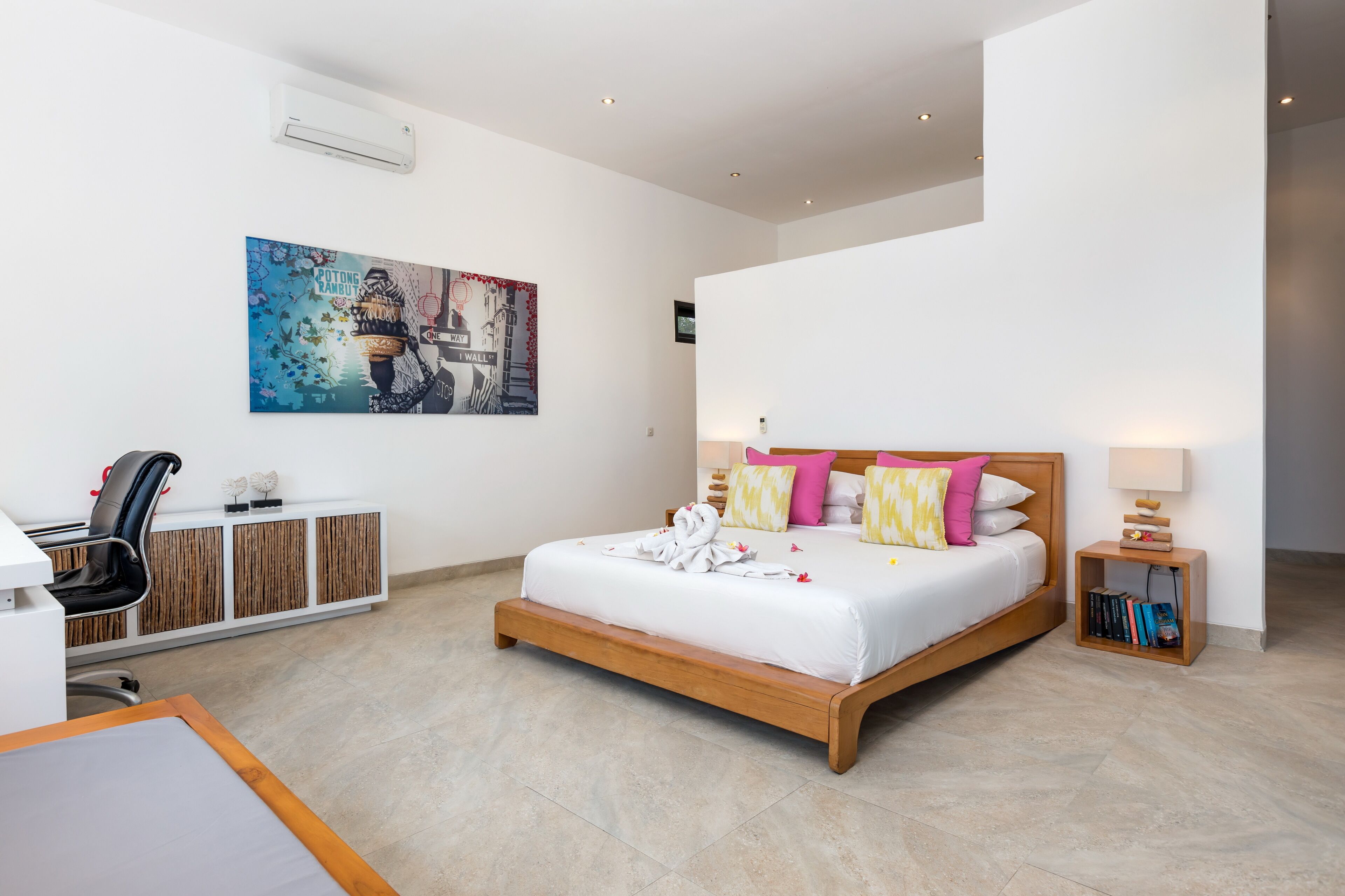 A modern bedroom with a wooden bed, white bedding, and colorful pillows. There is a desk with a chair and a bookshelf.