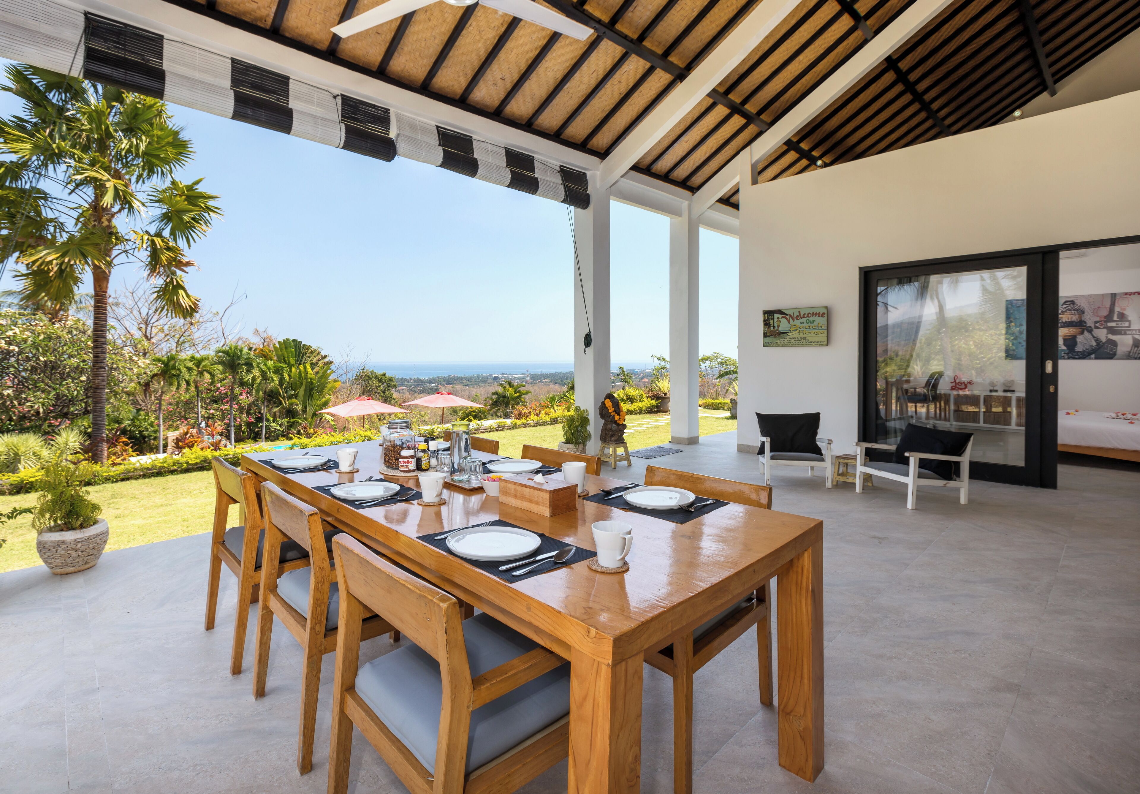 Un área de comedor al aire libre techada, con una mesa y sillas de madera, vista al océano y un jardín con diversas plantas.