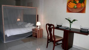 A bedroom with a four-poster bed, bedside table with lamp, wooden desk, chair, and a wall-mounted fruit basket.